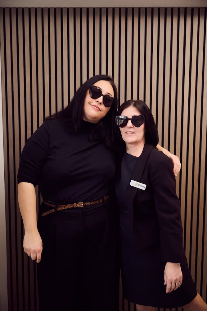Two women with dark hair wearing black outfits and heart-shaped sunglasses standing closely together in front of a wooden slat wall.