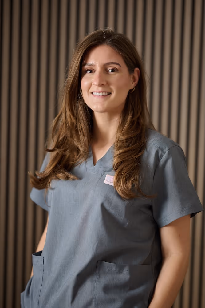 Smiling woman with long brown hair wearing gray medical scrubs standing against a striped background.