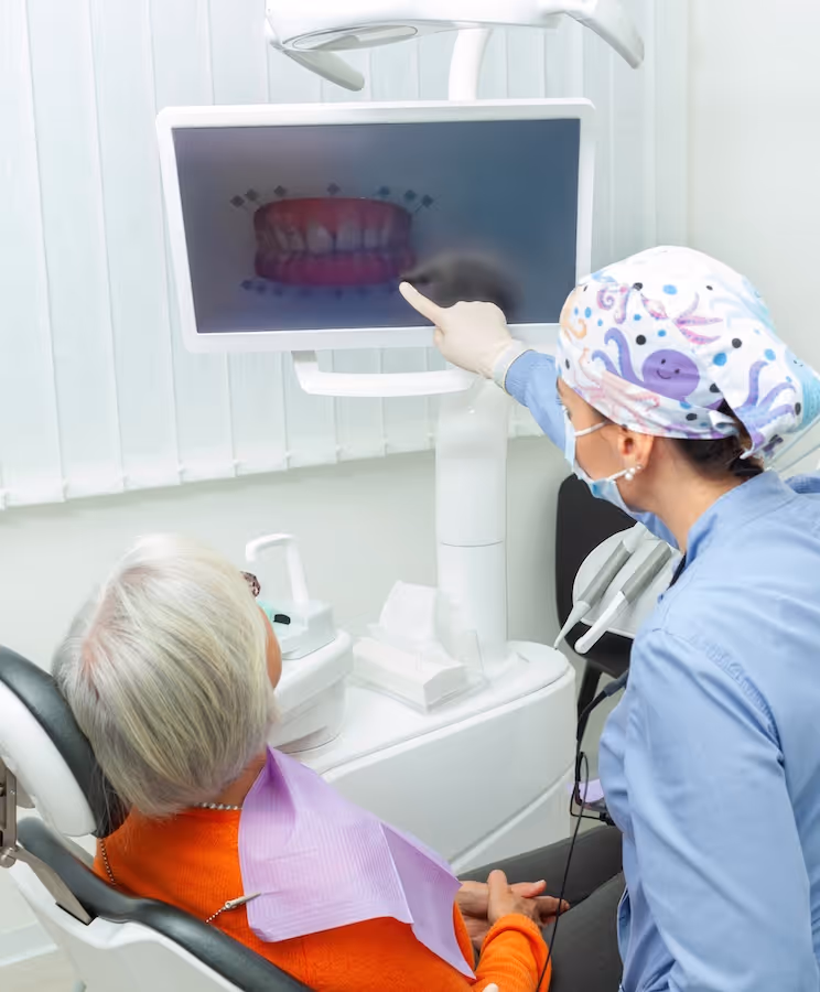 Dentist wearing gloves and a colorful cap pointing at a digital dental X-ray on a screen while an elderly patient in an orange sweater sits in the dental chair.