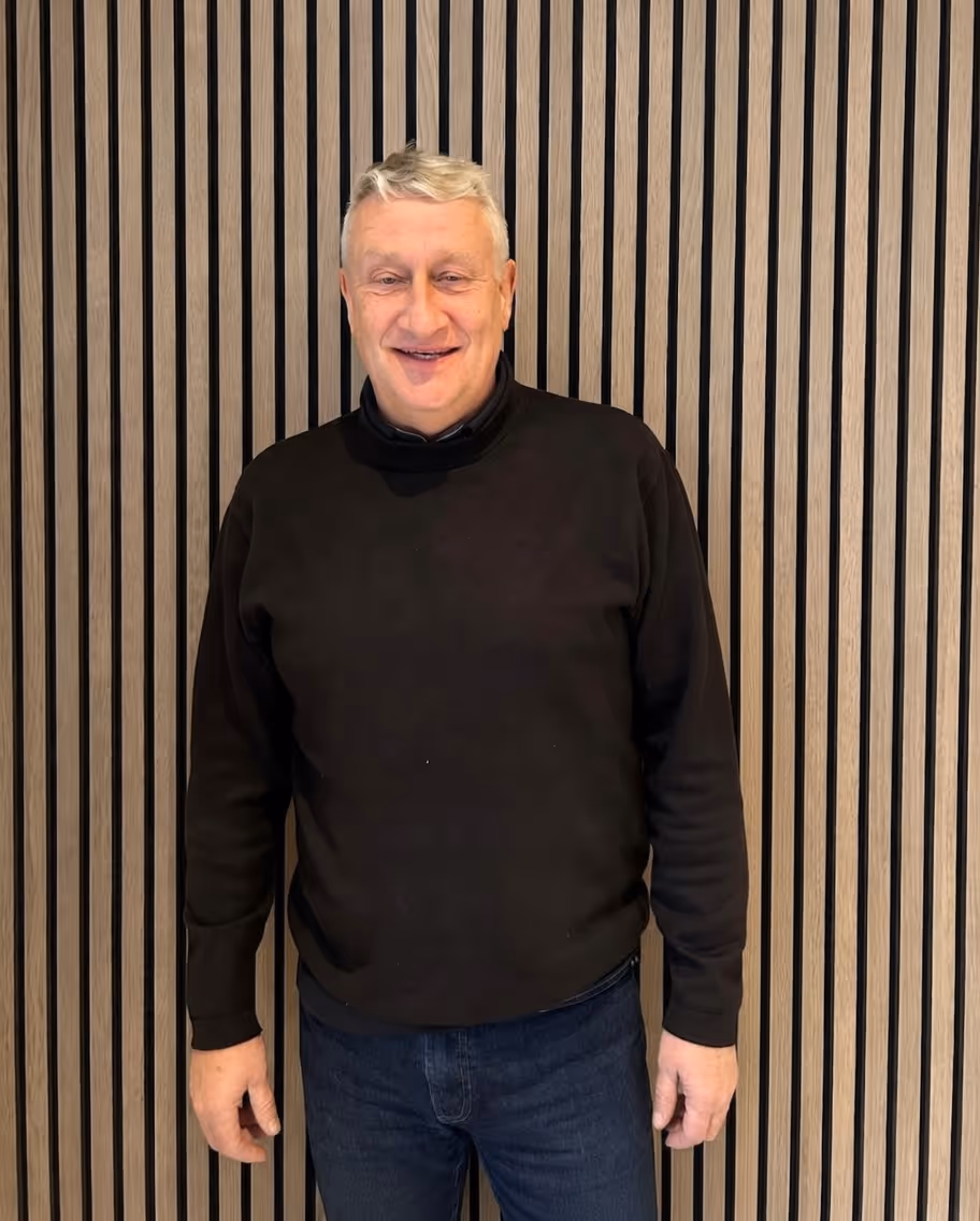 Smiling older man with short gray hair wearing a black sweater and blue jeans standing in front of a vertical wooden slat wall.