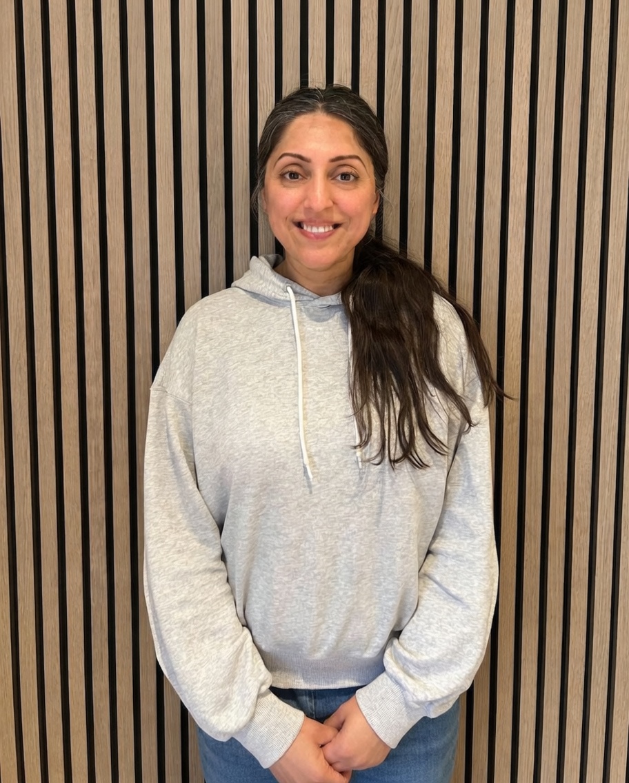 Smiling woman with long dark hair in a gray hoodie standing against a vertical striped wood panel wall.