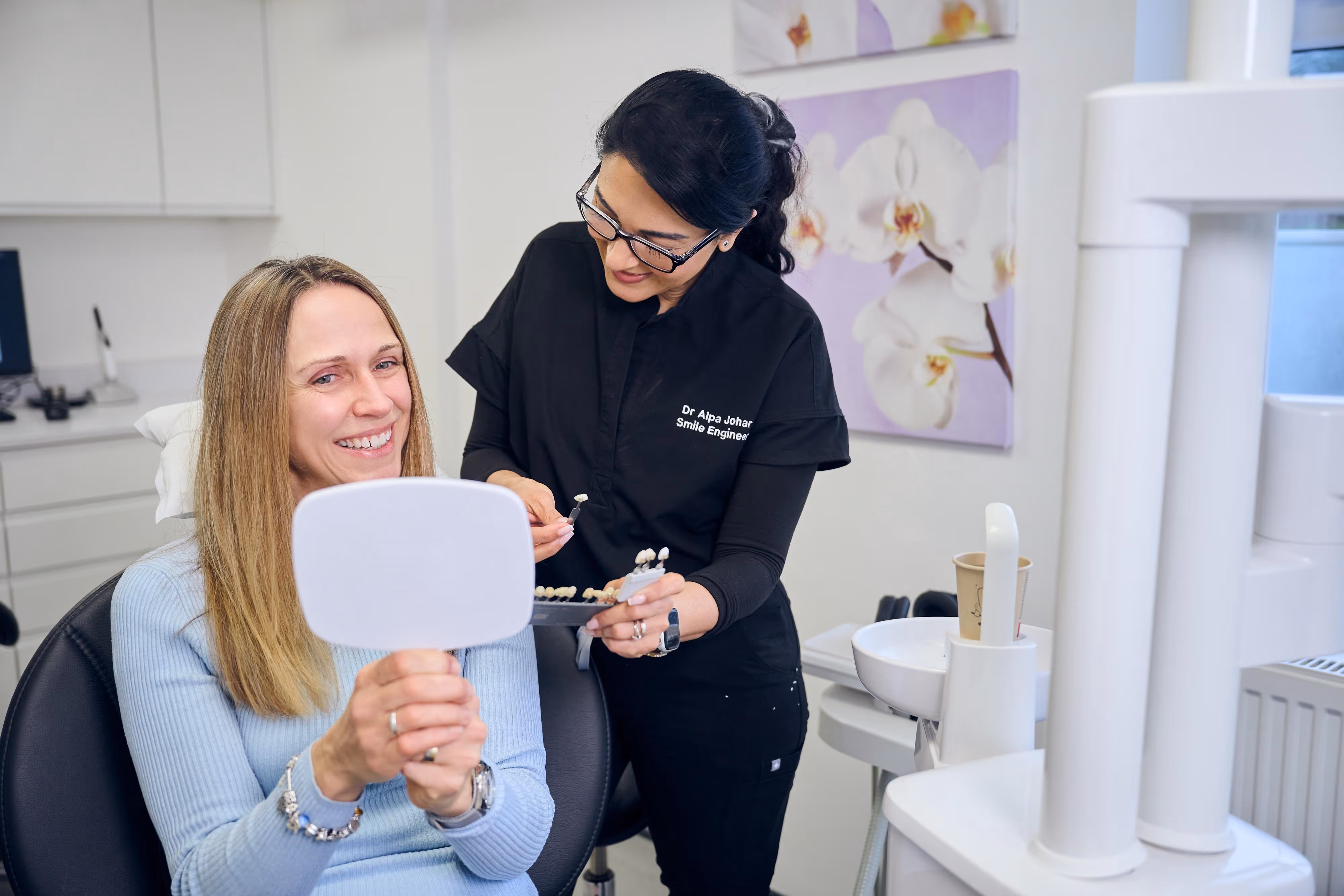 Dentist showing a female patient a dental shade guide while she smiles holding a handheld mirror in a dental clinic.