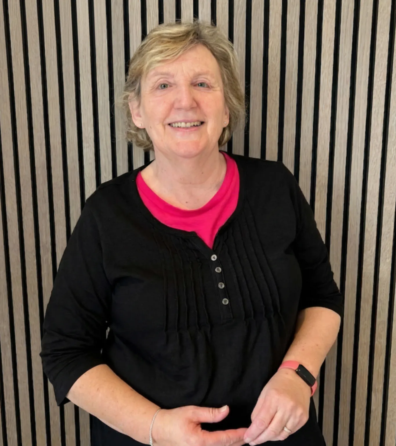 Smiling older woman with short blonde hair wearing a black top over a bright pink shirt, standing in front of a vertical striped wooden wall.