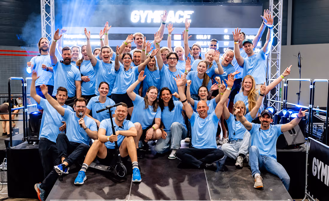 Group of smiling people in blue shirts waving hands on stage under a GymRace sign.