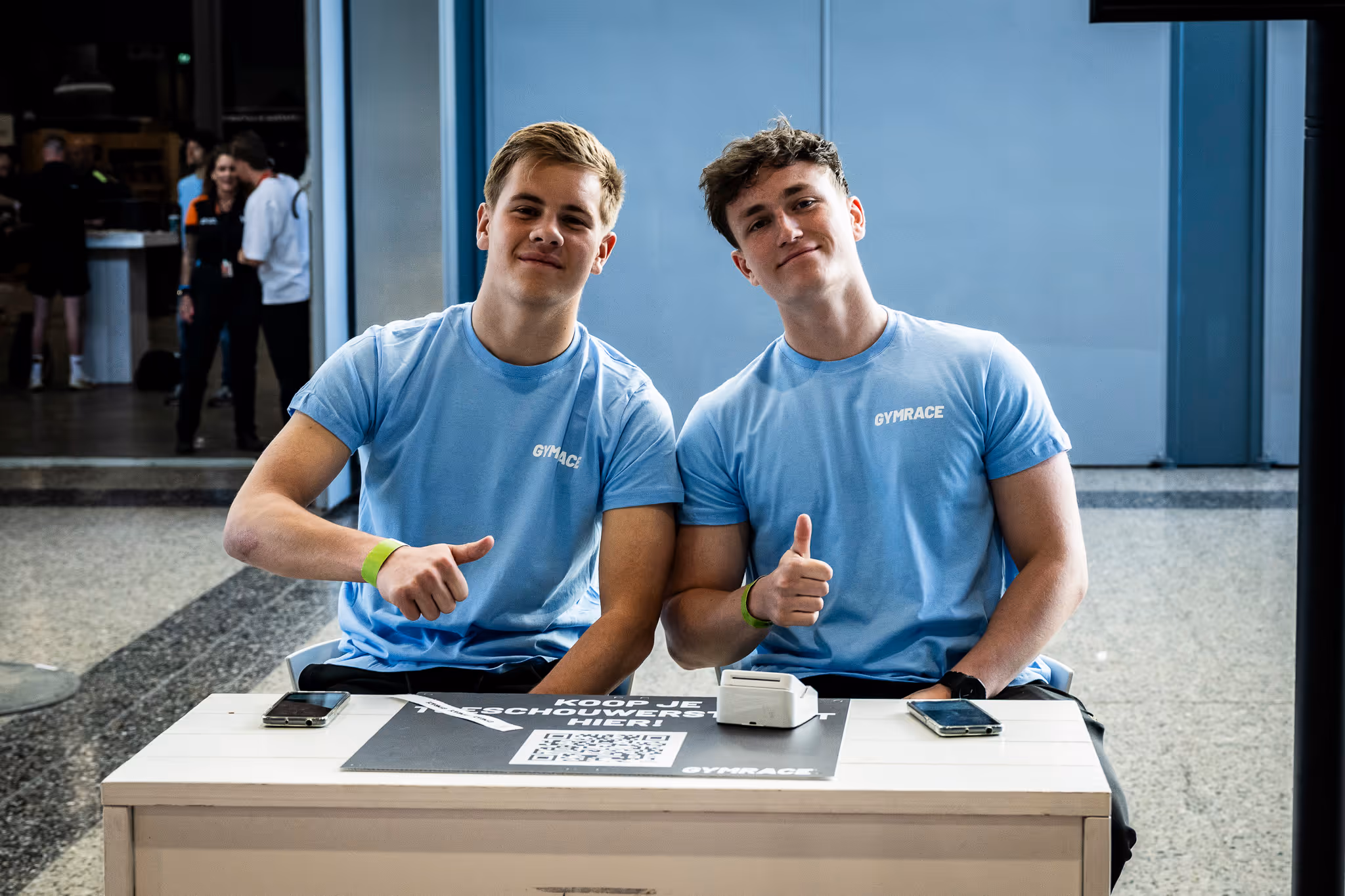 Two young men in light blue Gymrace t-shirts sitting at a table, giving thumbs up.