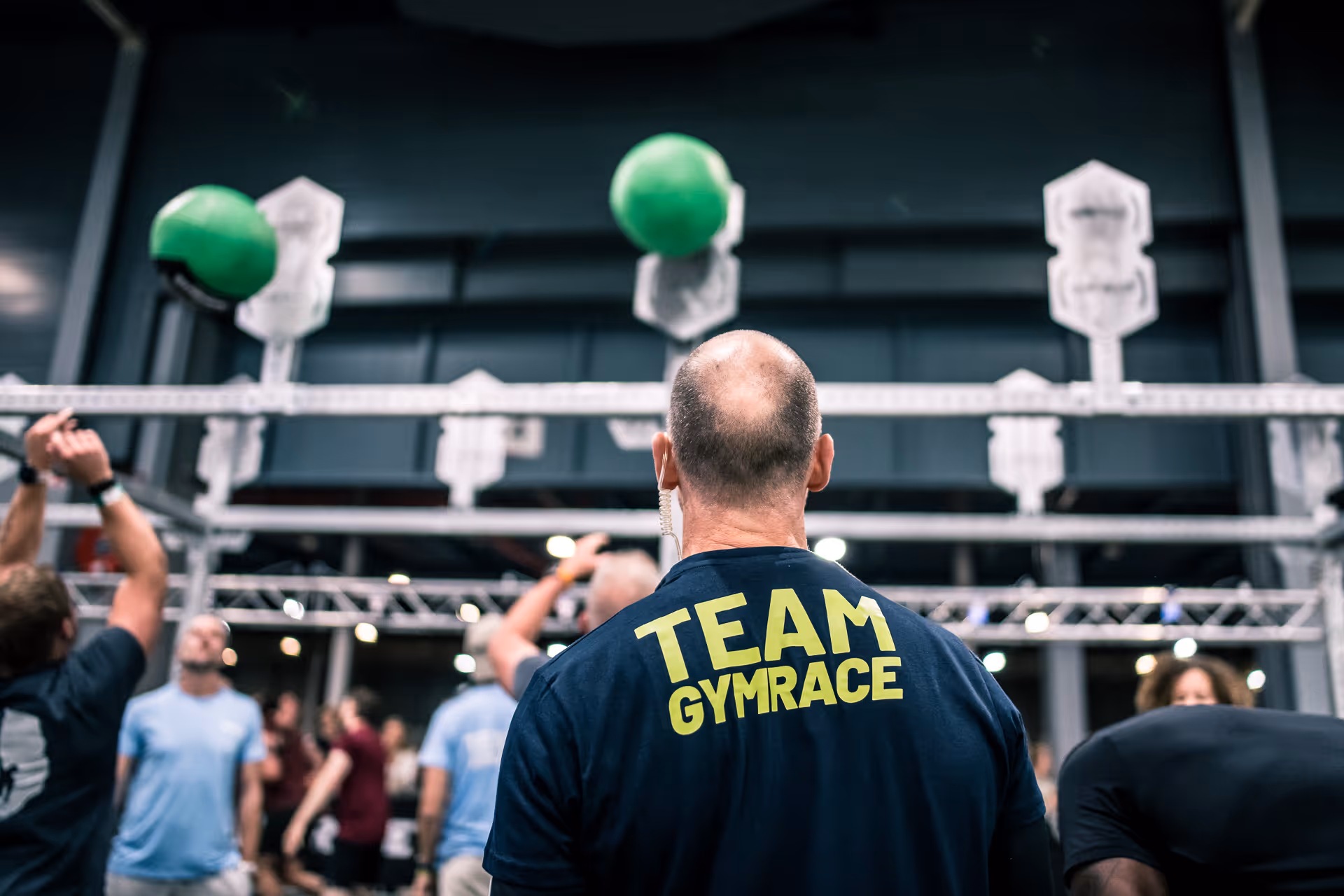 Bald man in Team GymRace shirt watches others throwing green balls at targets in gym competition.