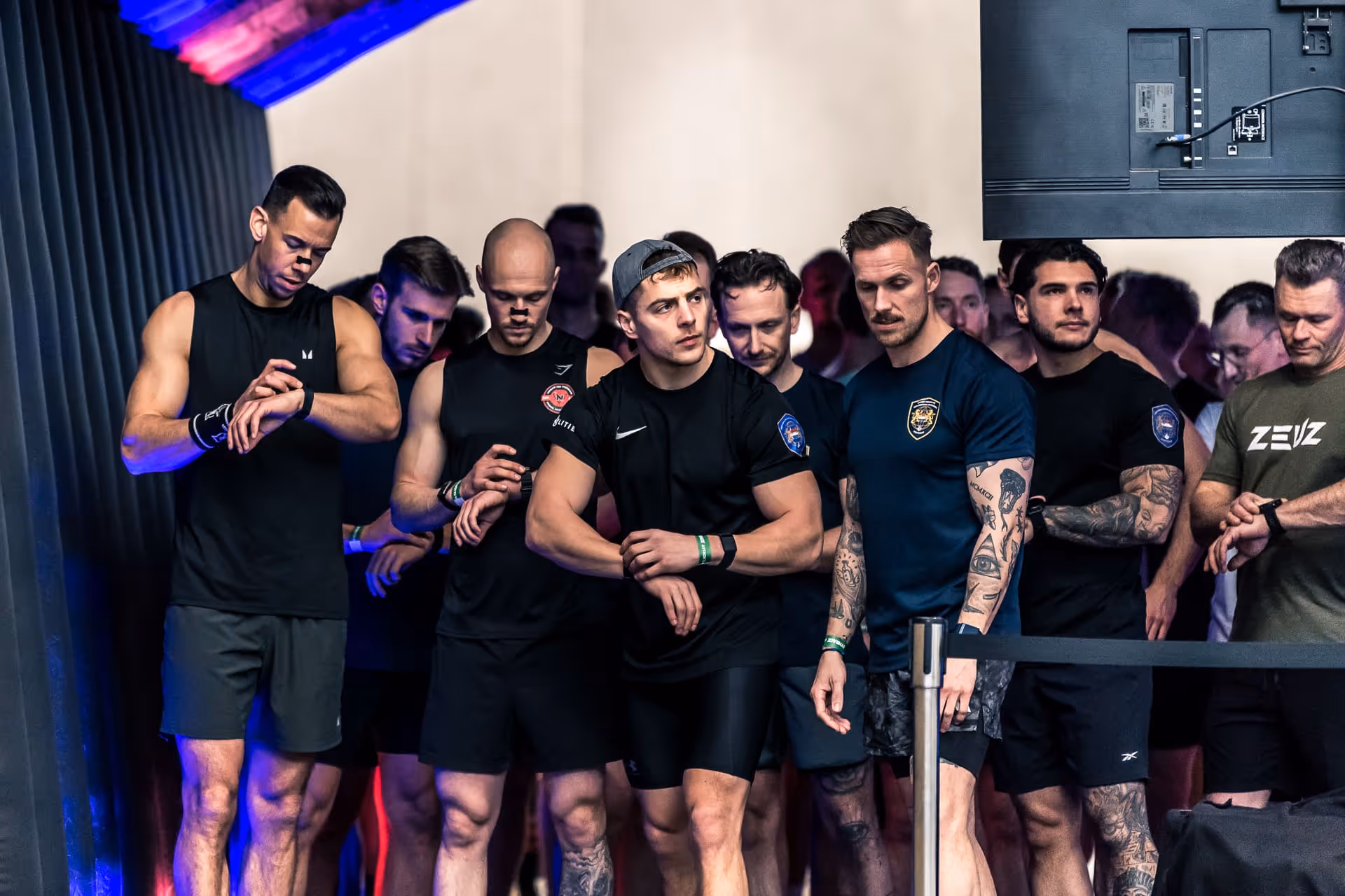 Group of muscular men in athletic wear standing in line, some checking their watches or fitness bands.
