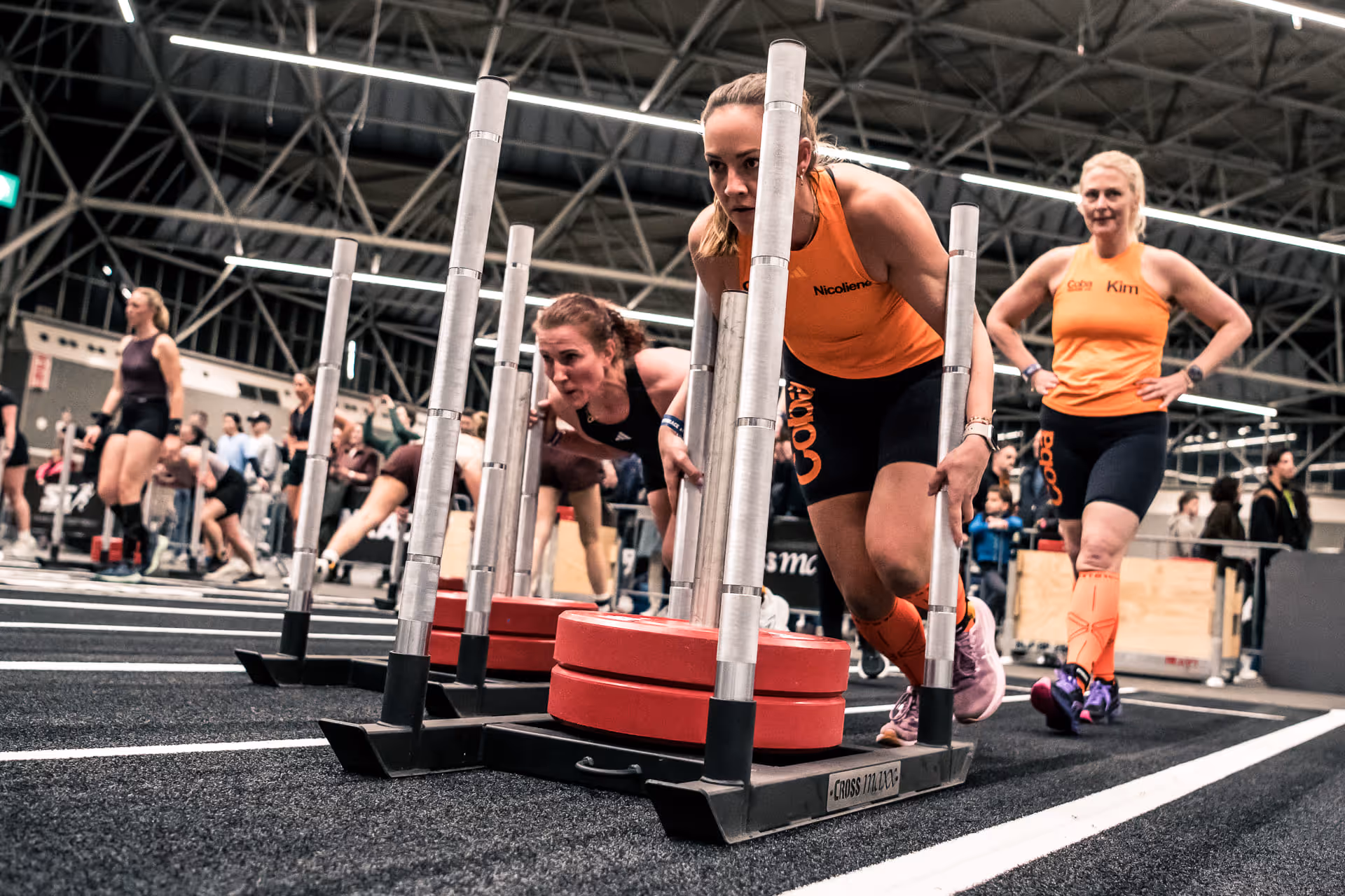 Women competing in sled push strength event on black gym floor with spectators in background.