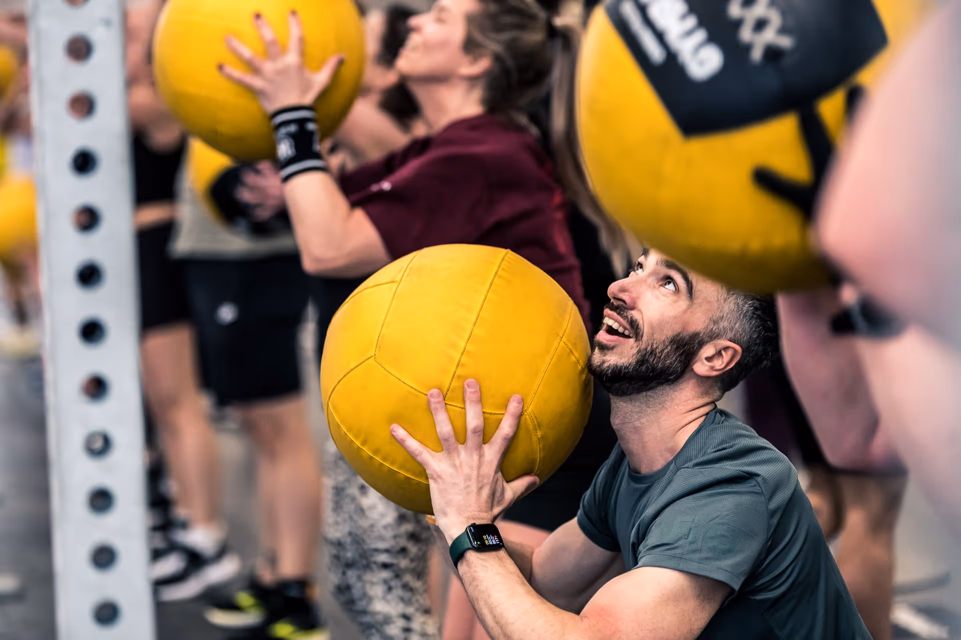 Man and others exercising with yellow medicine balls against a gym wall in a fitness class.