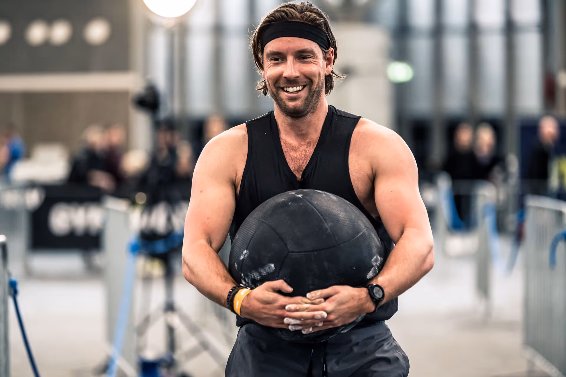 Smiling man in black tank top holding a black slam ball in a gym or sports event setting.