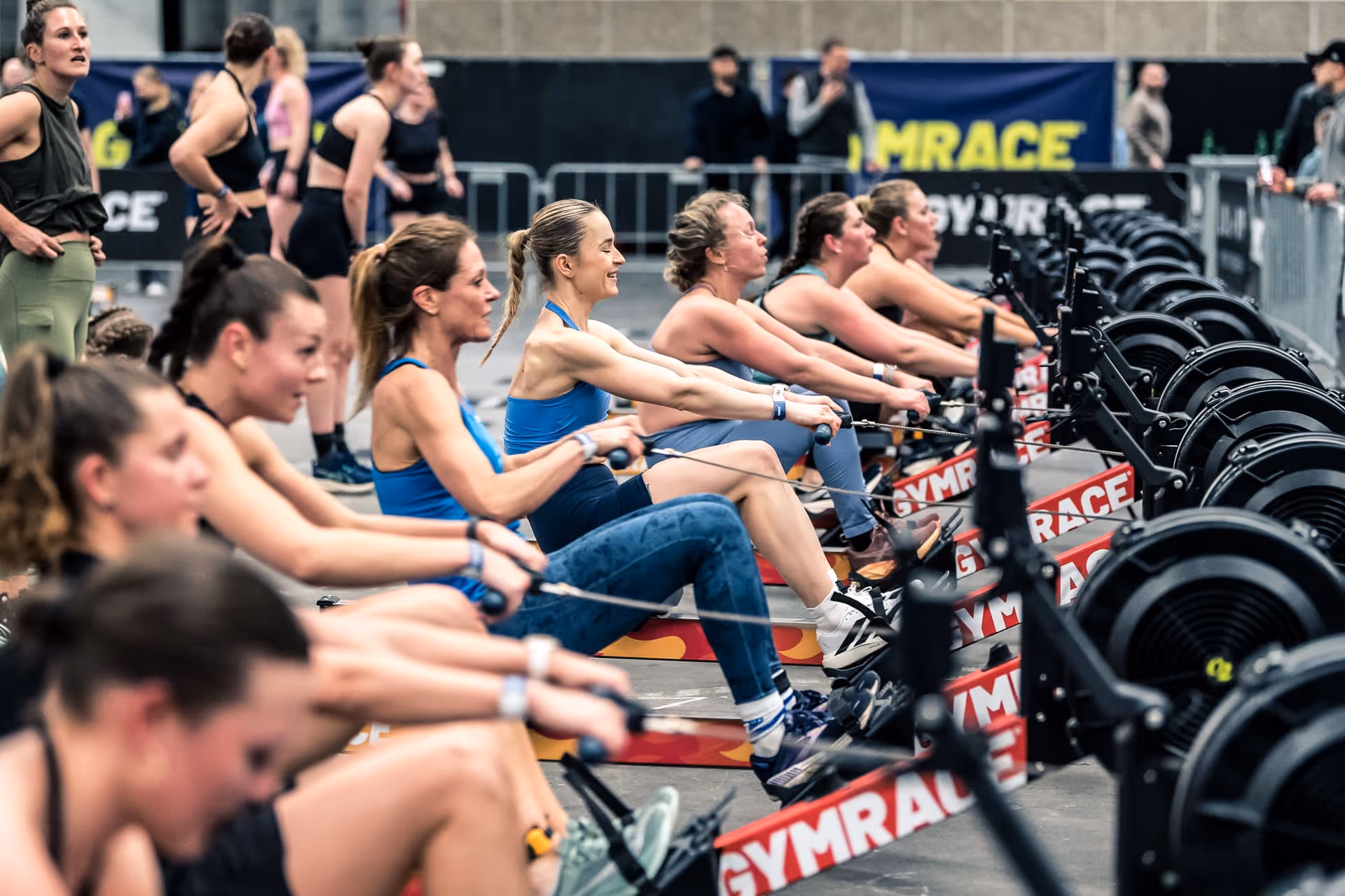Women competing on rowing machines in an indoor fitness event with spectators in the background.