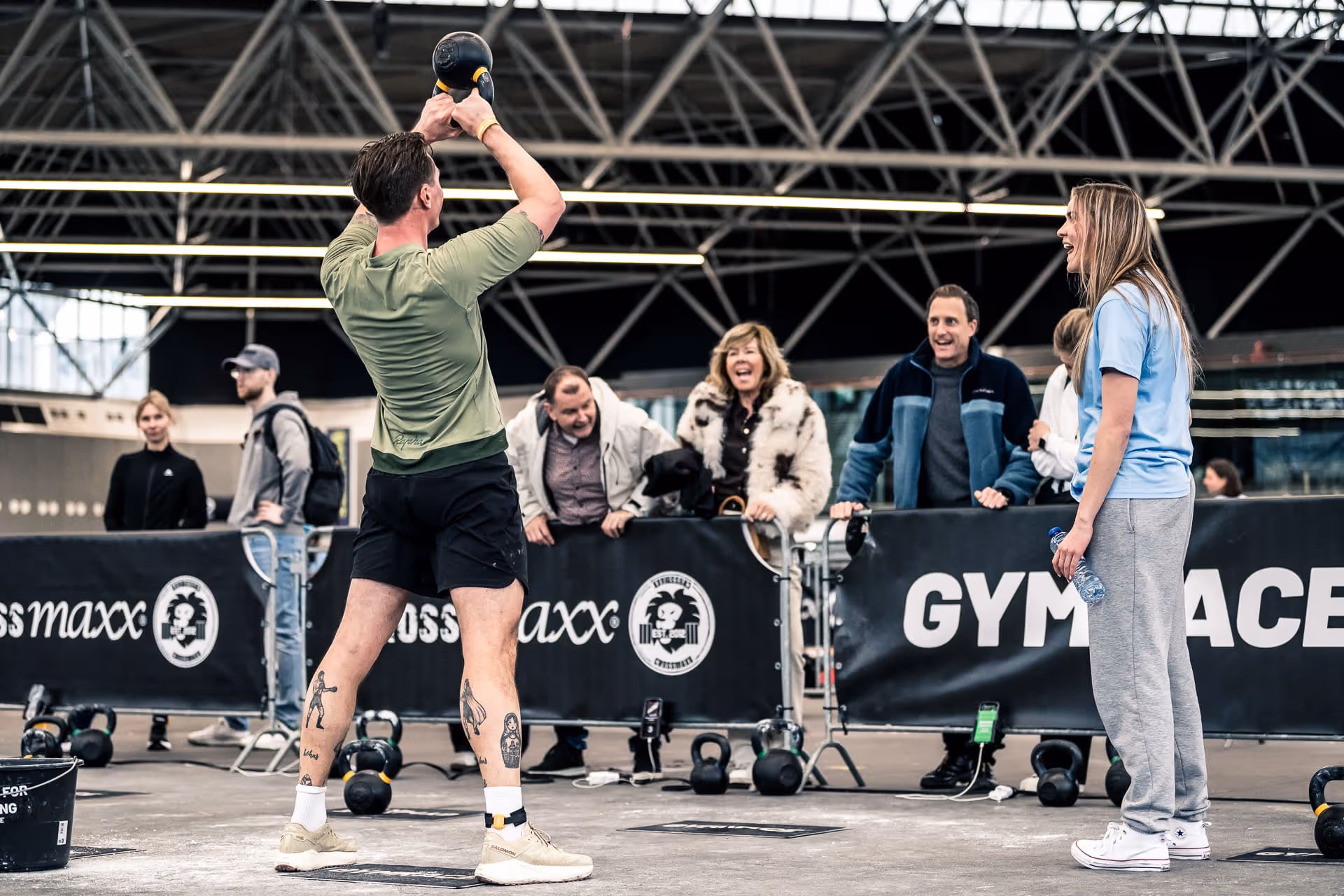 Man lifting kettlebell overhead during workout as a woman watches and crowd cheers behind barriers.