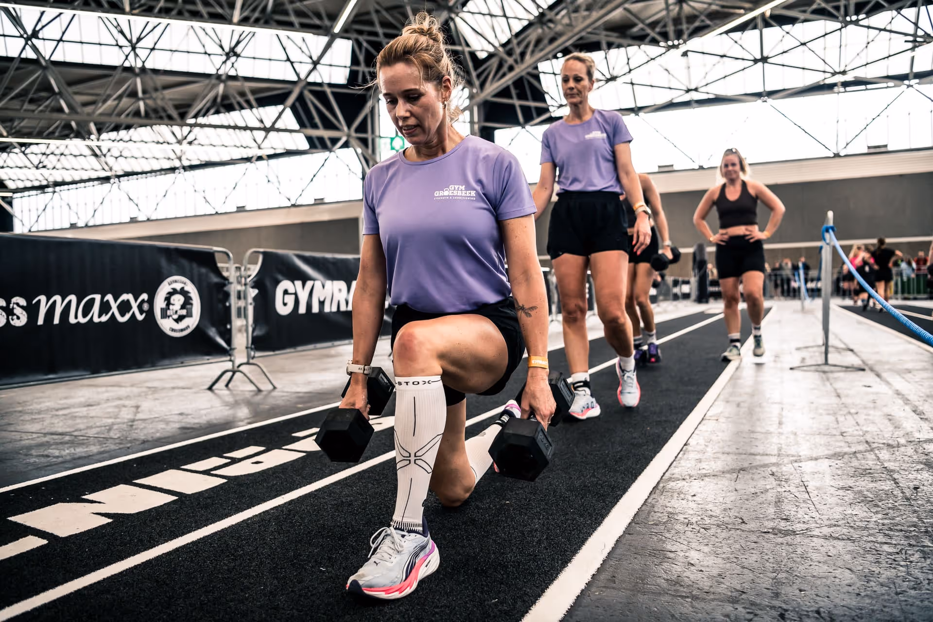 Women in purple shirts exercising with dumbbell lunges indoors on a black track.