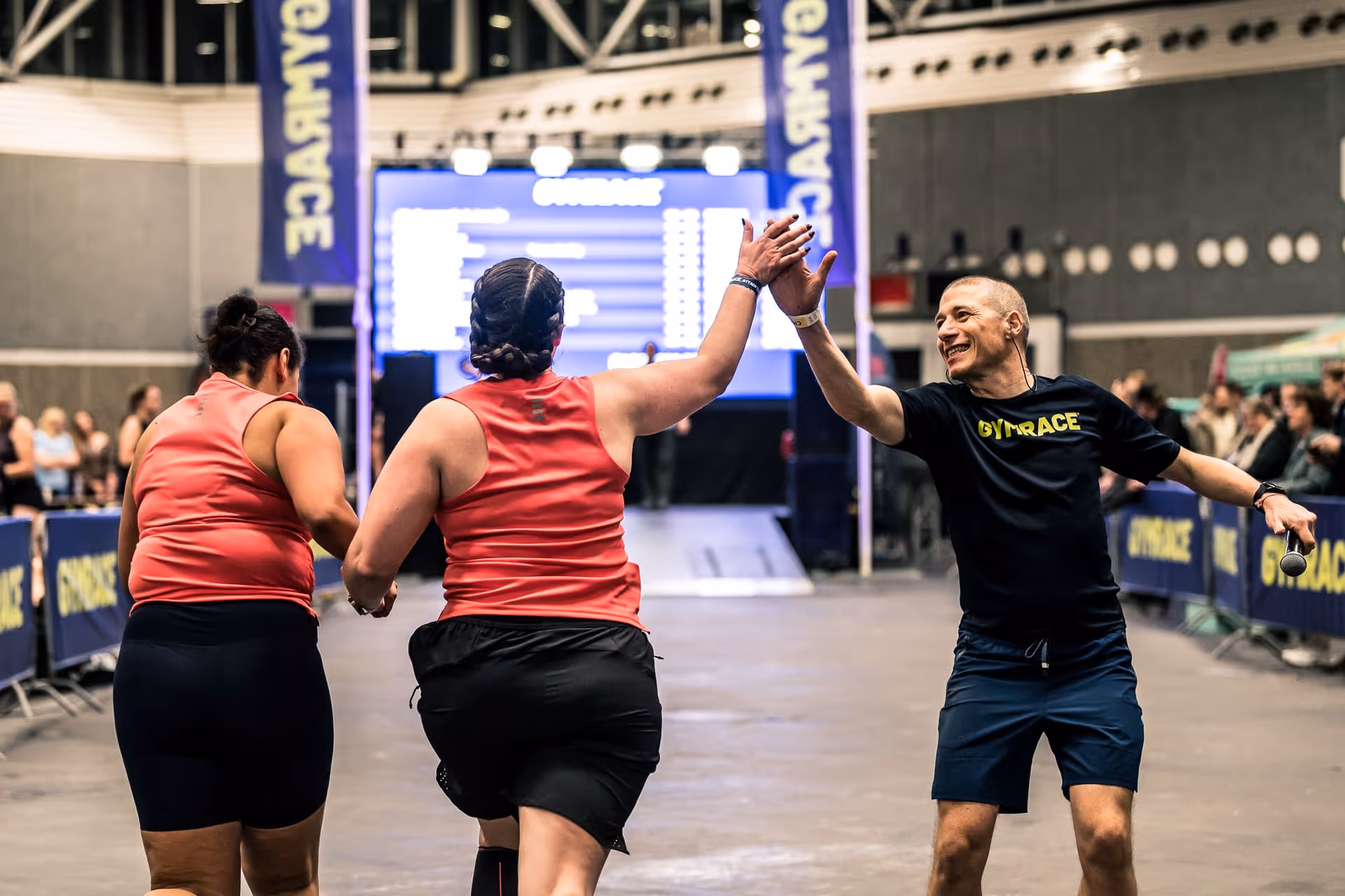Two participants in red shirts high-five a man in a GymRace shirt at an indoor race finish.