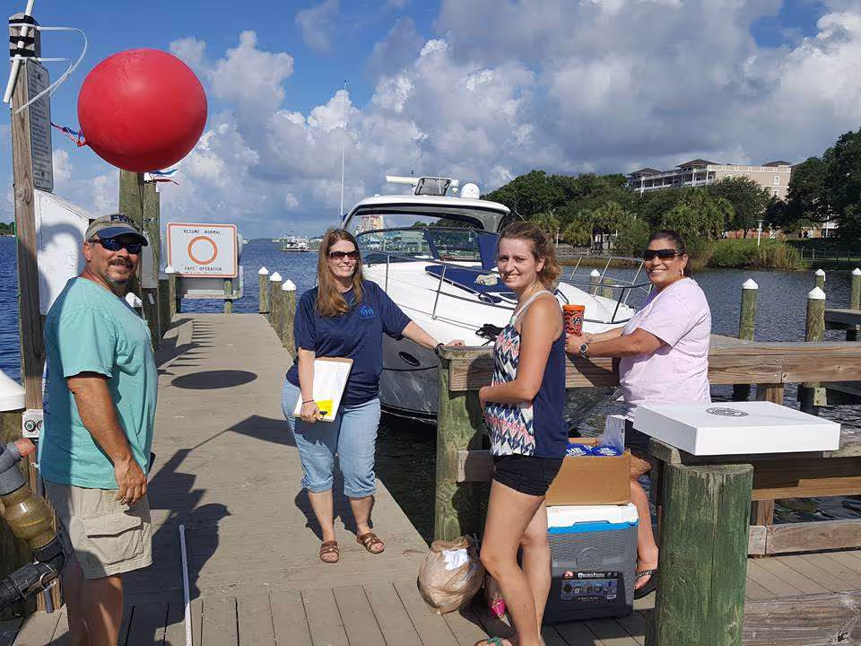 Four people standing on a dock next to a boat on a sunny day at a marina.