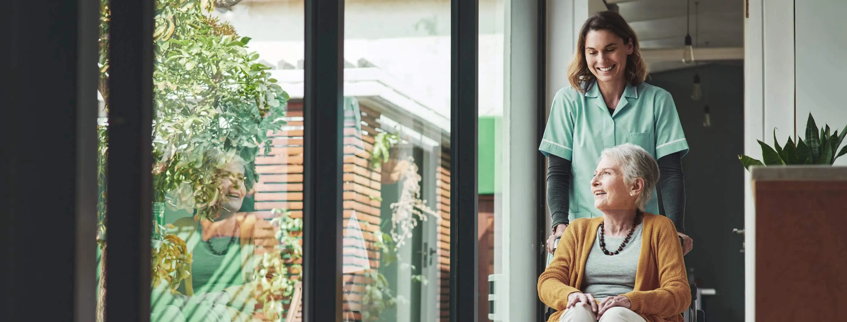 Lächelnde Pflegekraft schiebt eine ältere Frau im Rollstuhl durch helle Innenräume mit großen Fenstern – professionelle ambulante Pflege und liebevolle Seniorenbetreuung im Alltag.
