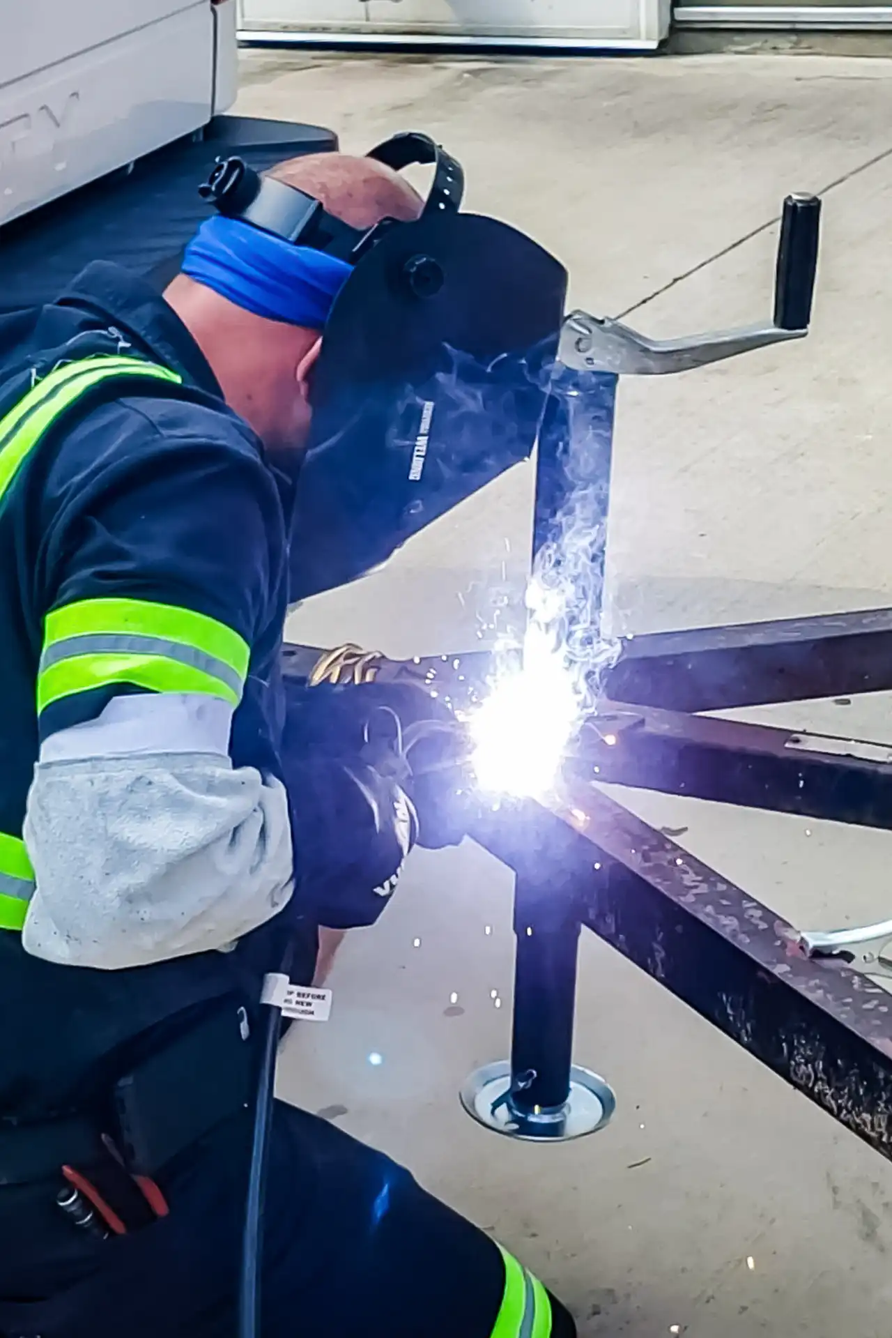 Welding sparks erupt as mechanic fuses trailer jack leg to frame; “WELDING ARMOUR” text visible on dark face shield