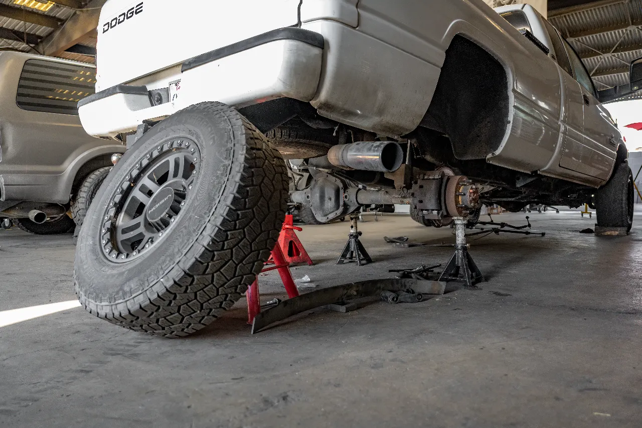 Rear suspension repair on a Dodge truck lifted on jack stands, axle exposed with leaf spring removed and DODGE tailgate visible.