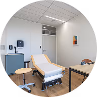 Clean medical examination room with an exam table, rolling stool, small refrigerator, and a desk with a chair.
