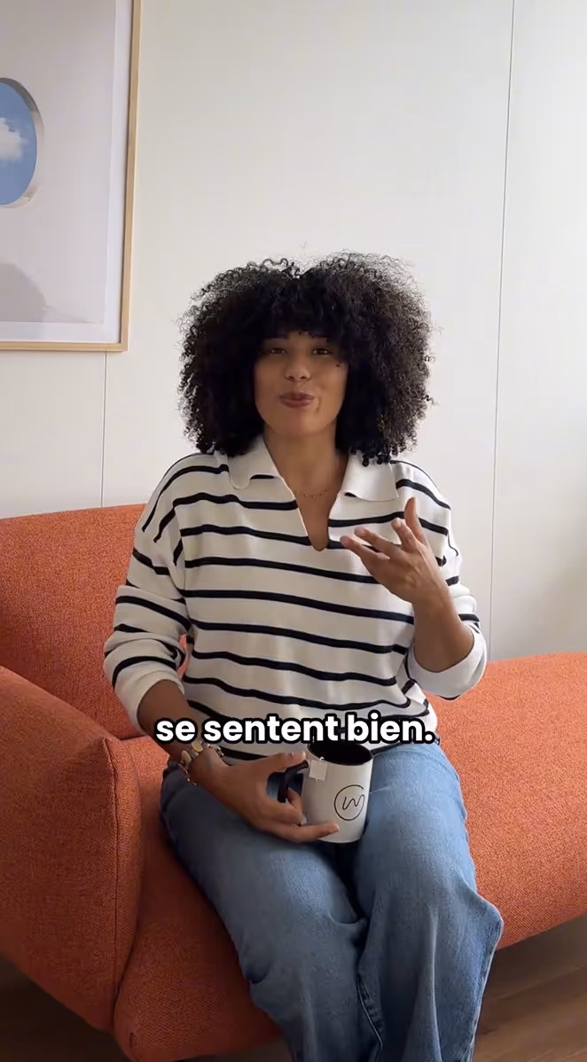 Woman with curly hair sitting on orange chair holding a mug, wearing a white and black striped shirt and blue jeans.