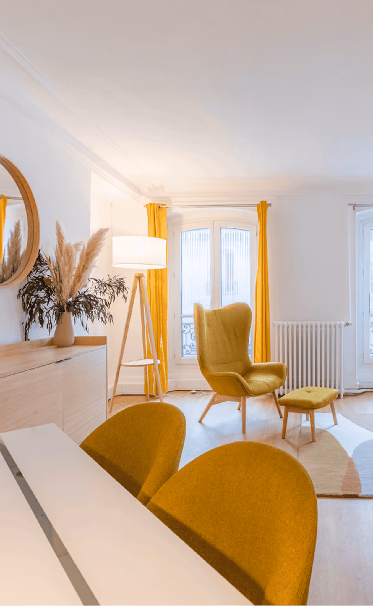 Bright modern living room with mustard yellow armchair, matching ottoman and chairs, wooden sideboard, floor lamp, and round wall mirror.
