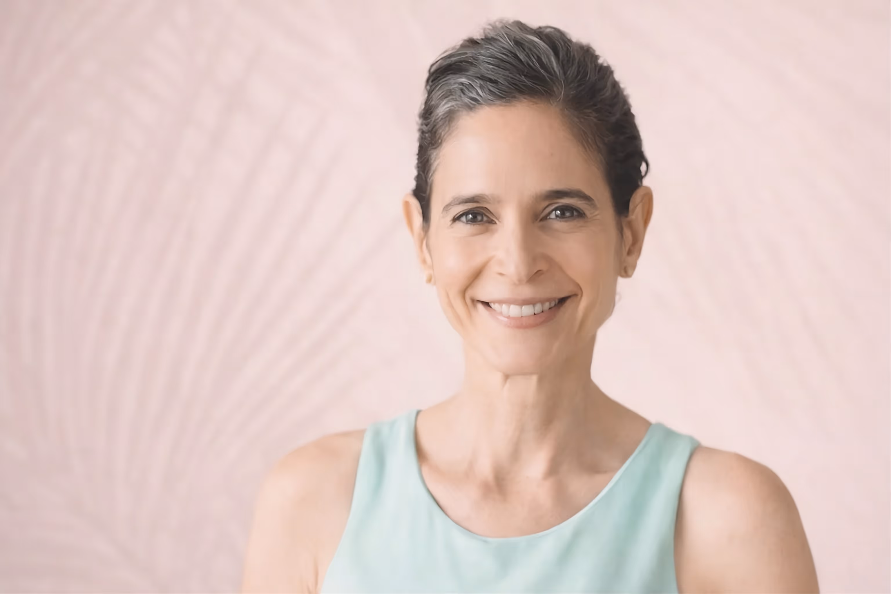 Smiling middle-aged woman with short gray hair wearing a light blue sleeveless top, against a pale pink background.