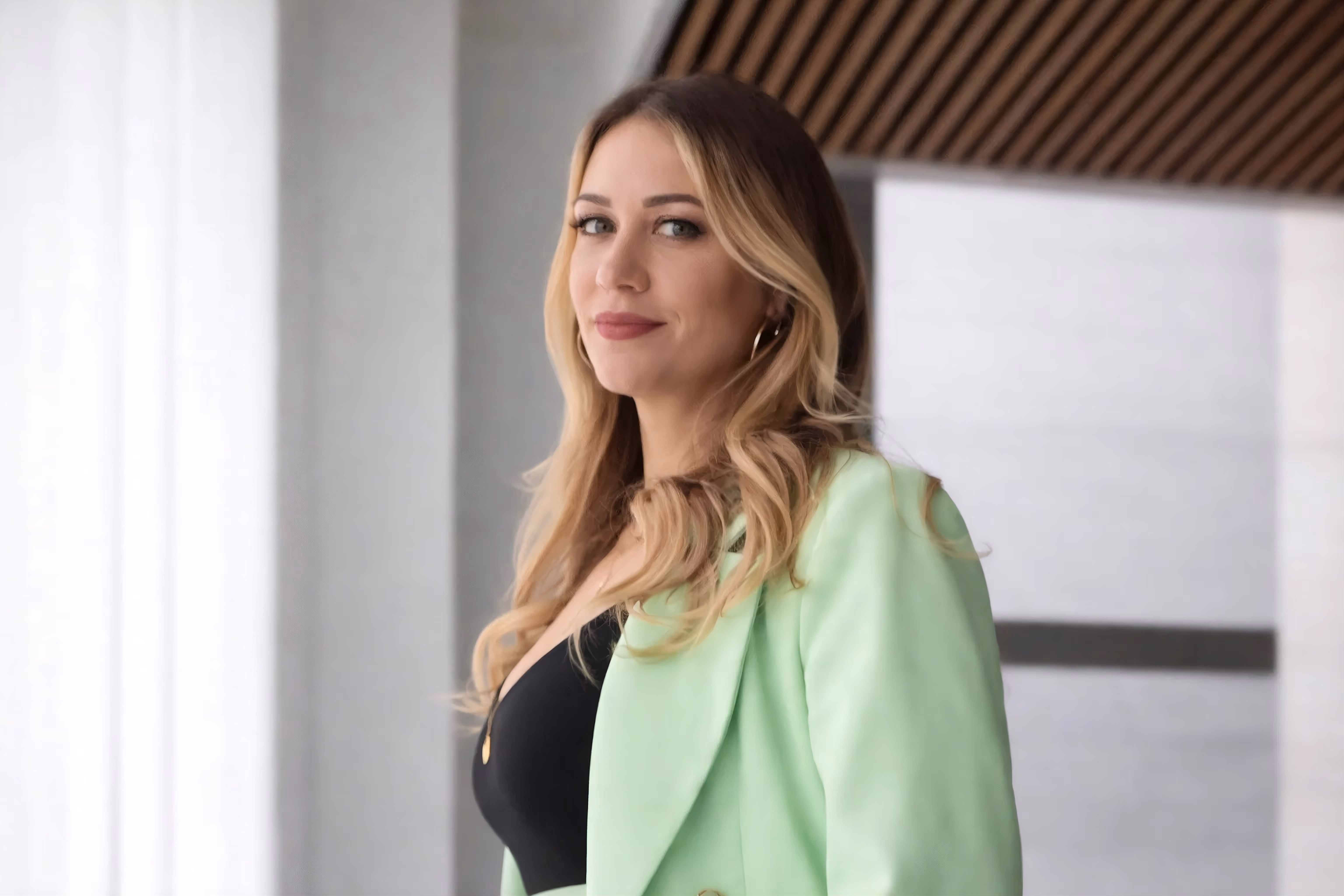 Blonde woman wearing a light green blazer and black top looking to the side in a modern indoor setting.