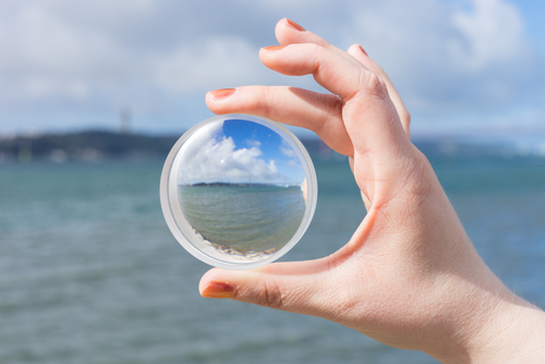 Hand holding a clear glass sphere reflecting a seascape with sky, clouds, and distant land.