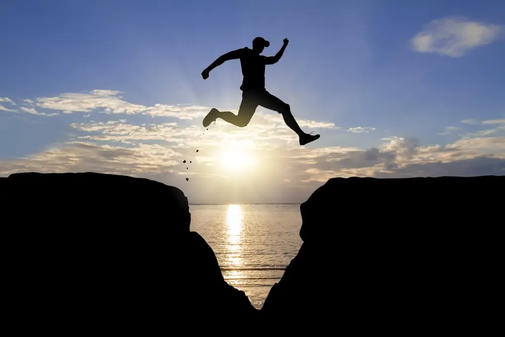Silhouette of a man jumping over a gap between two cliffs with the sun setting over the ocean in the background.
