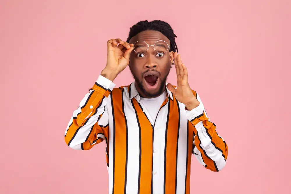 Surprised man with glasses wide-eyed and mouth open against a pink background.
