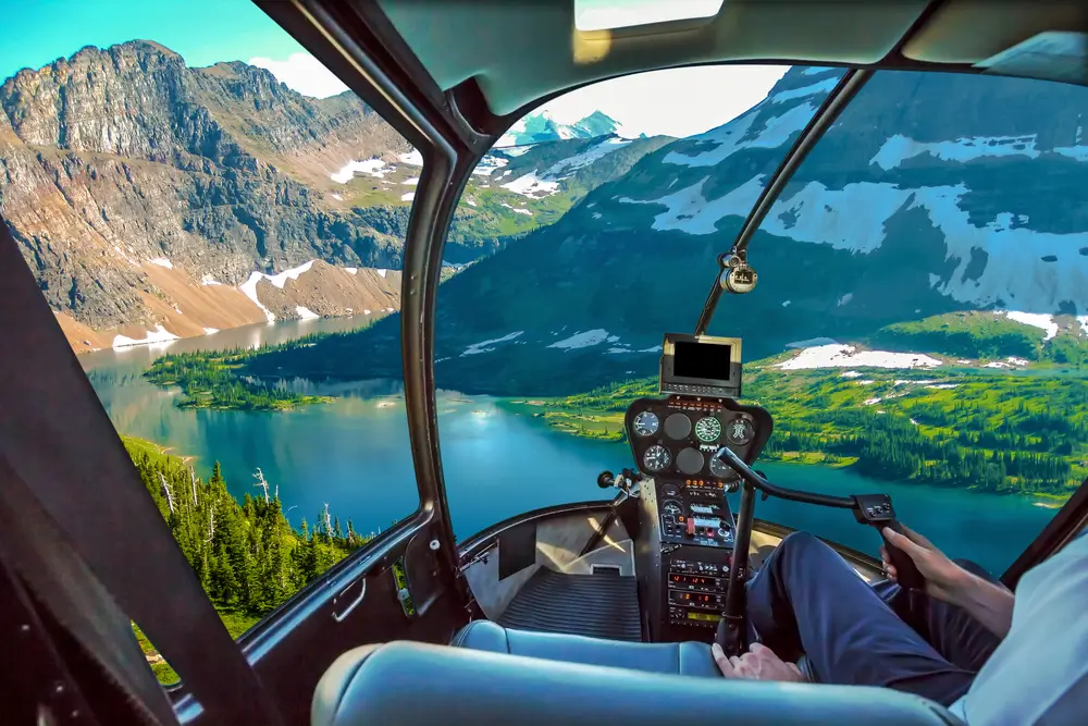 View from inside a helicopter cockpit overlooking a turquoise lake surrounded by mountains with patches of snow and green forests.
