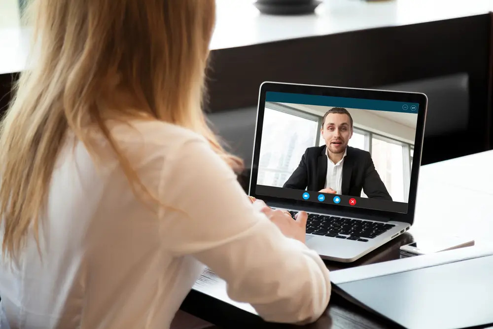 Woman with long hair in a white blouse participating in a video call with a man in a suit on a laptop.