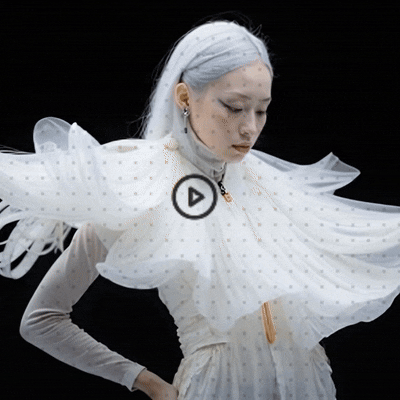Cinematic GIF of a woman with long silver hair wearing an ethereal white gown, with a play button overlay. Surreal fashion and modern digital design.