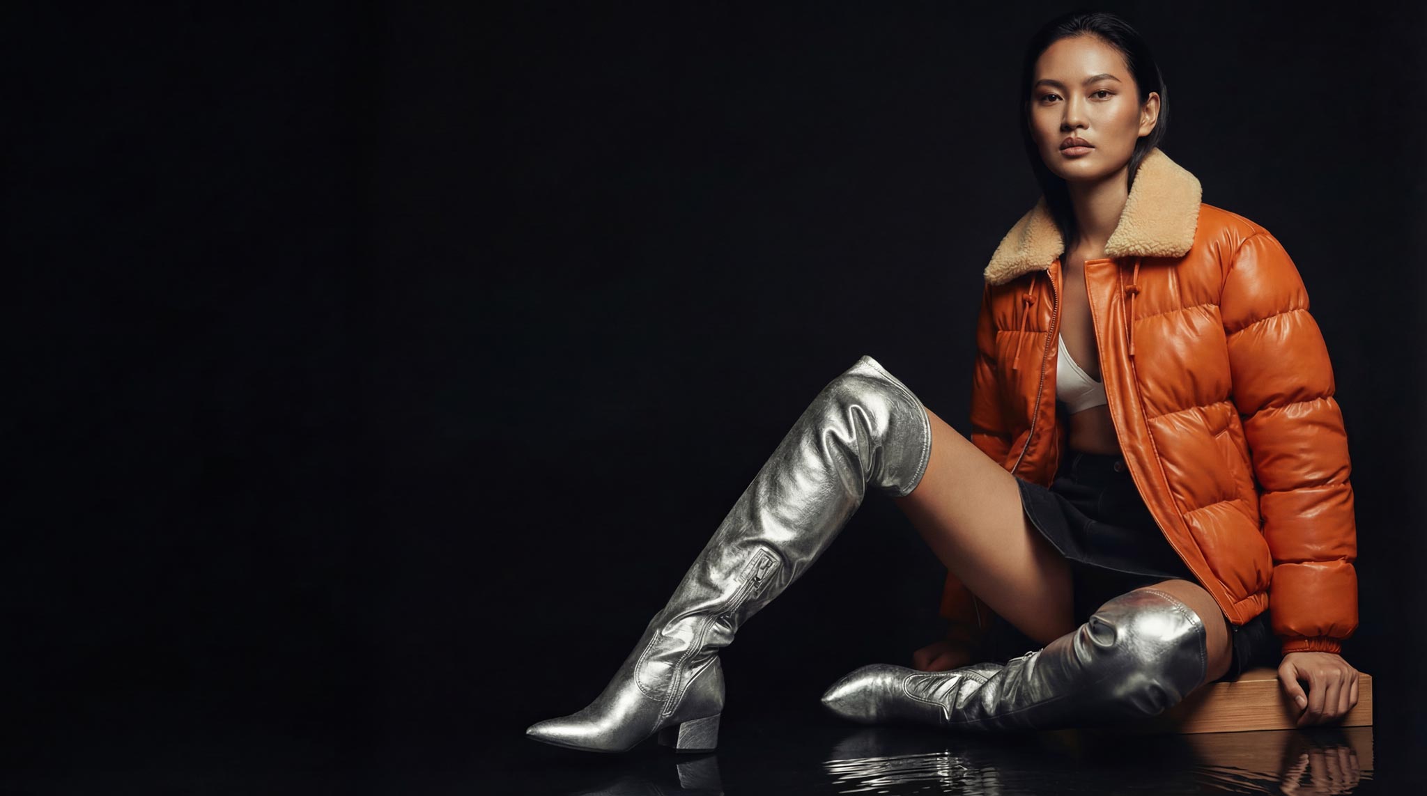 Fashion model sitting on a wooden block wearing a shiny orange puffer jacket with a beige fur collar, silver thigh-high boots, a white top, and a black skirt against a black background. Made with The Fabricant Intelligent Tools