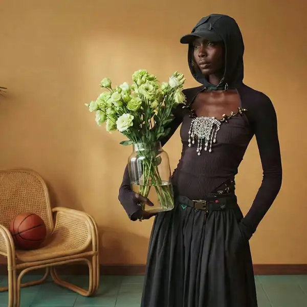 Fashionable person in a hooded top and cap holding a glass vase with green flowers, standing beside a wicker chair with a basketball. Made with The Fabricant Image-To-Video tool