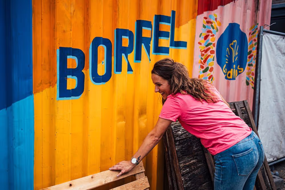 Vrouw in roze shirt leunt en lacht tegen een kleurrijke container met het woord 'BORREL' erop.