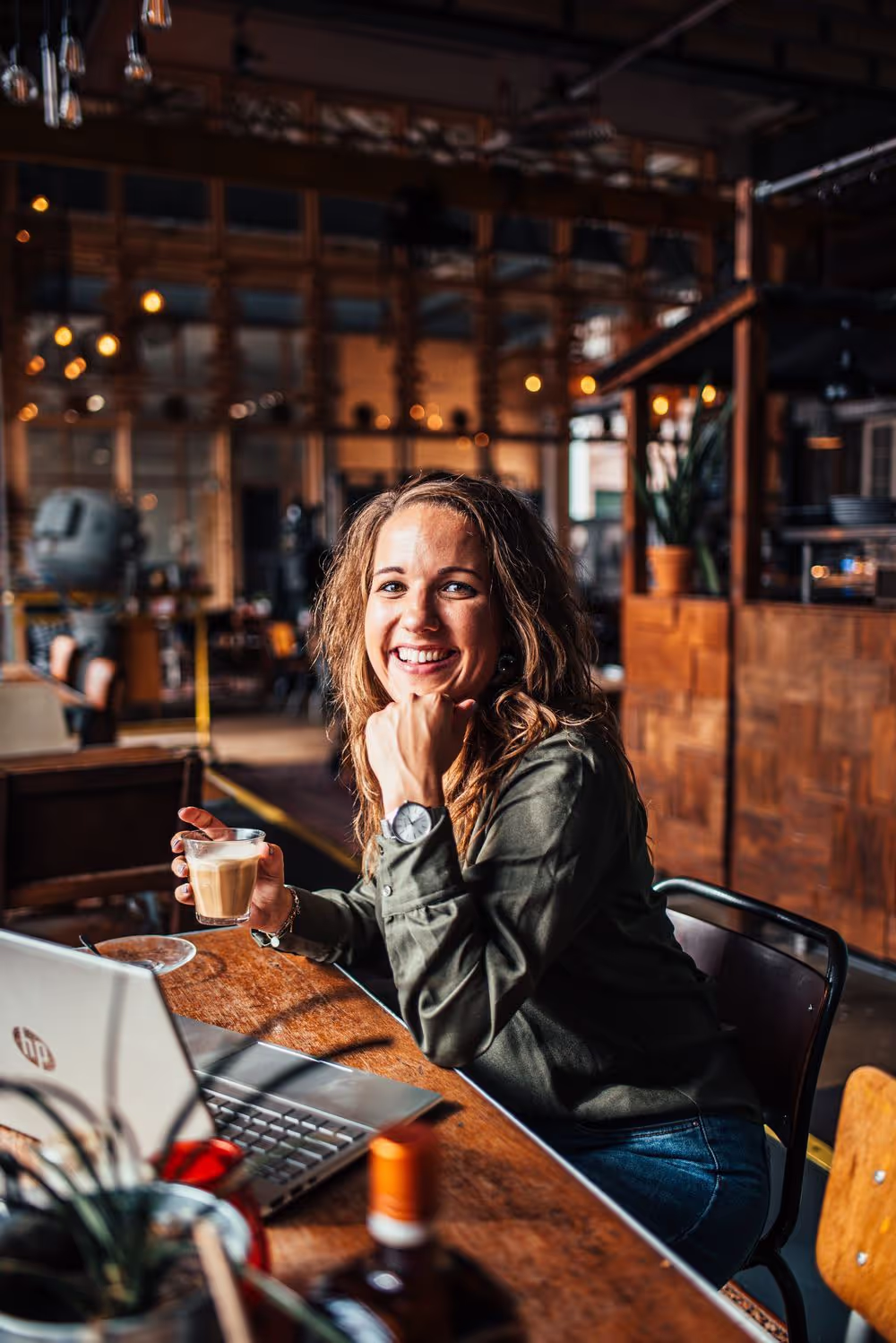 Vrouw die glimlacht terwijl ze in een café aan een tafel met een laptop en een kopje koffie zit.