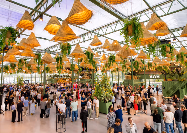Drukke evenementhal met veel mensen, grote plantenbakken en hangende lampen van gevlochten materiaal, met een bar aan de zijkant.