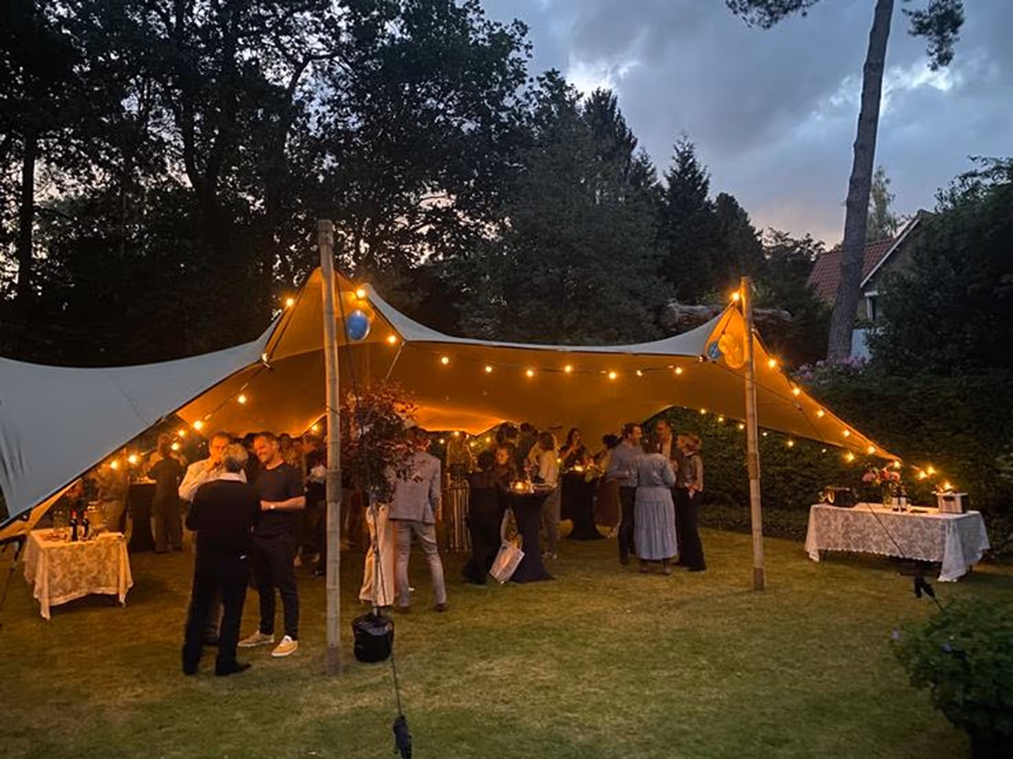 Gezelschap van mensen geniet van een avondfeest onder een verlichte stretchtent in een tuin.