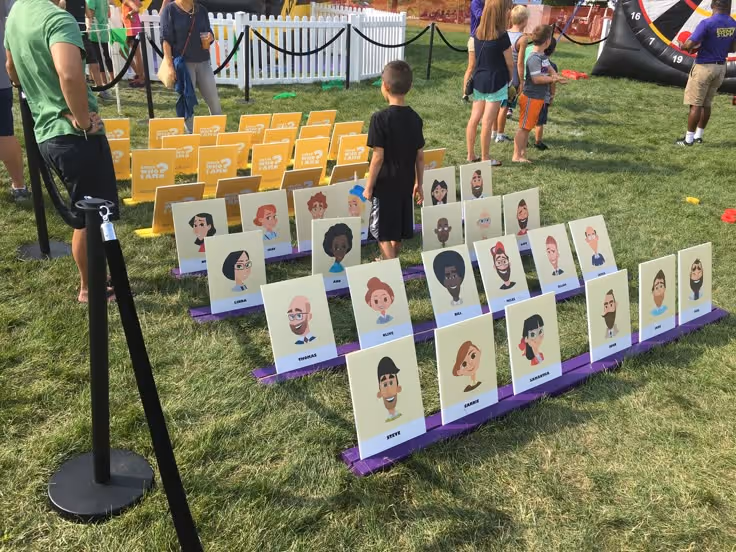 Buitenactiviteit met levensgrote kaartjes met cartoonportretten van mensen op gras; kinderen en volwassenen bekijken de opstelling.