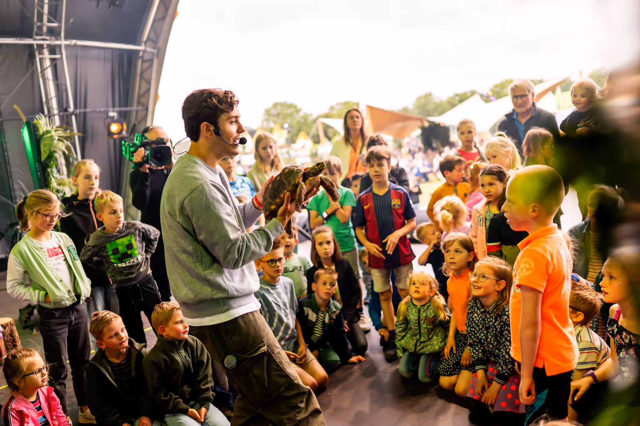 Een man met een microfoon toont een schildpad aan een groep kinderen die aandachtig toekijken.