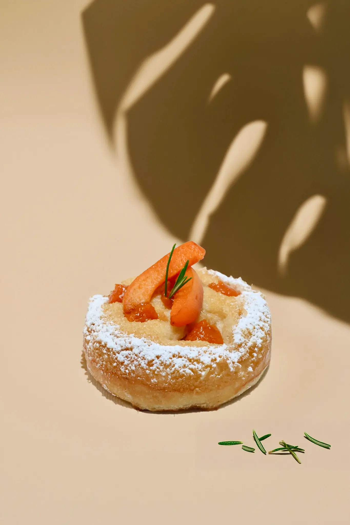 La viennoiserie du mois aux abricots et romarin sur fond beige avec une ombre de feuille et des brindilles de romarins éparpillées