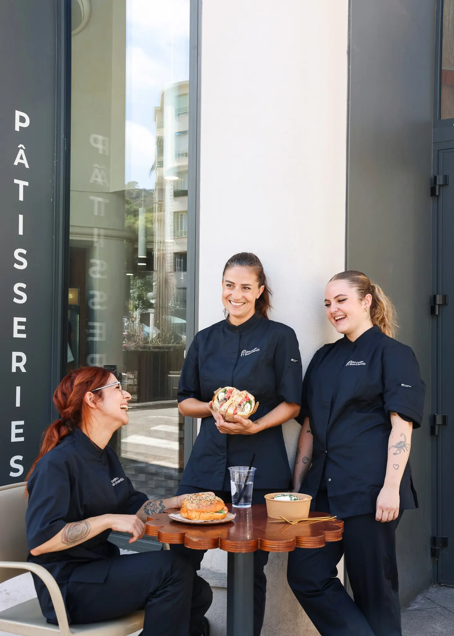 Notre chef cuisinière ainsi que ses deux commis autour d’une table en train de rigoler entre elles, sont posés devant elles des salades et sandwichs Les petits marchands