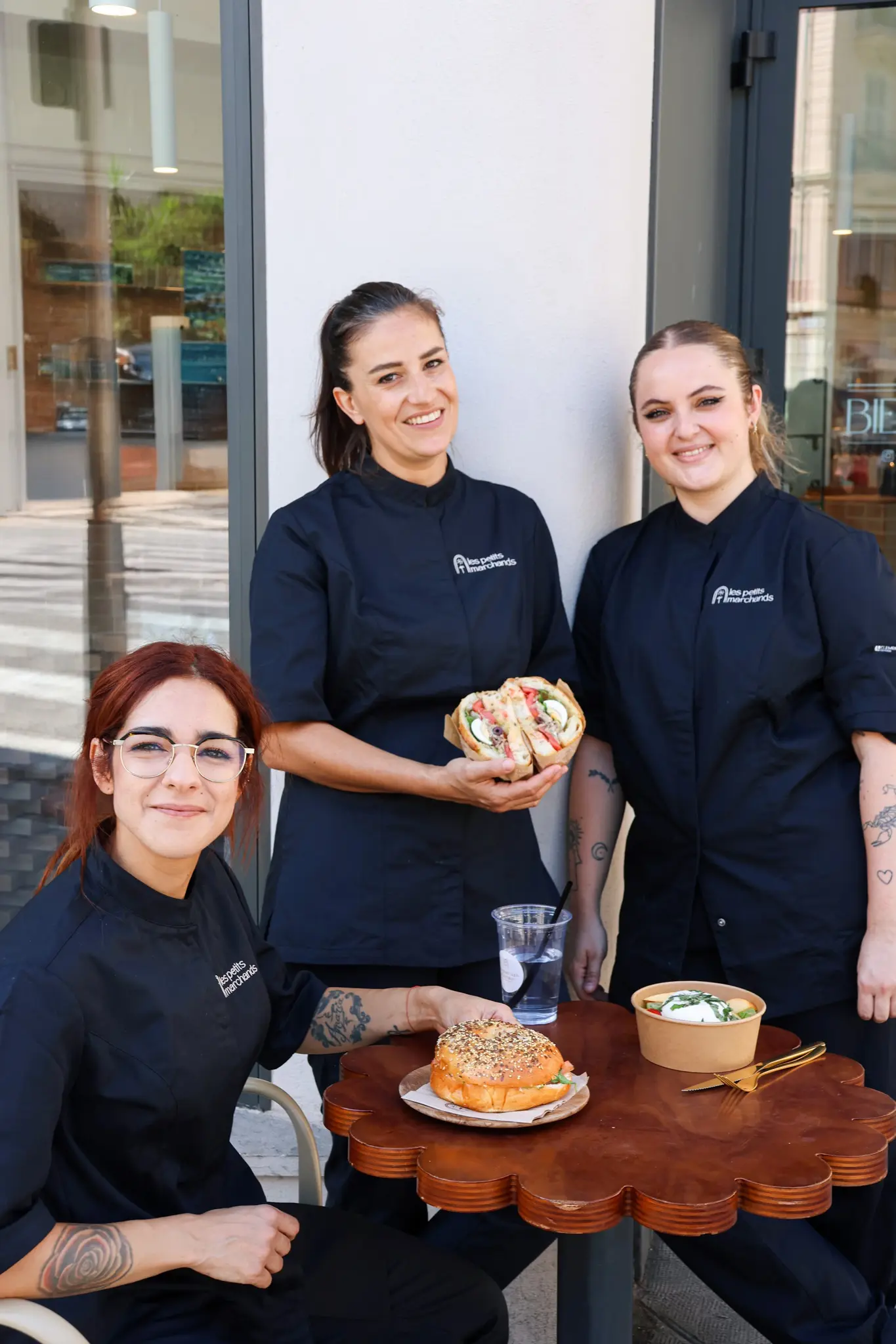 La cheffe cuisinière accompagnée de ses secondes, autour d’une table Les Petits Marchands, portant des sandwichs et salades, regardant la caméra en souriant