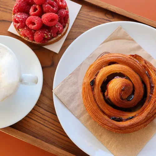 Un plateau en bois avec une tarte framboise individuelle, un pain au raisin sur une serviette et un cappuccino