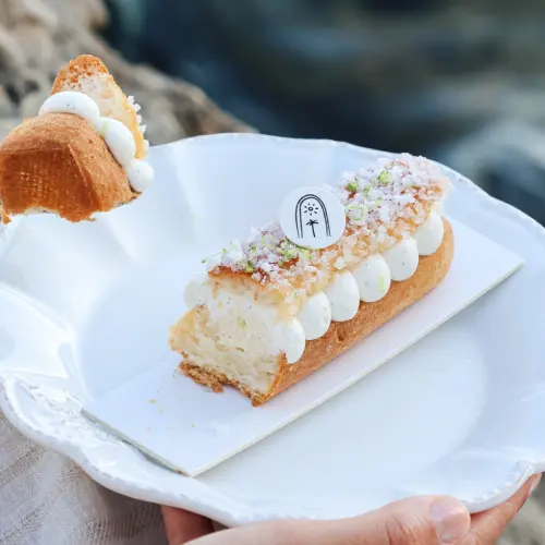  ⁠⁠Une personne coupe un morceau de la tropézienne fleur d’oranger sur une assiette blanche avec une cuillère, face à la mer