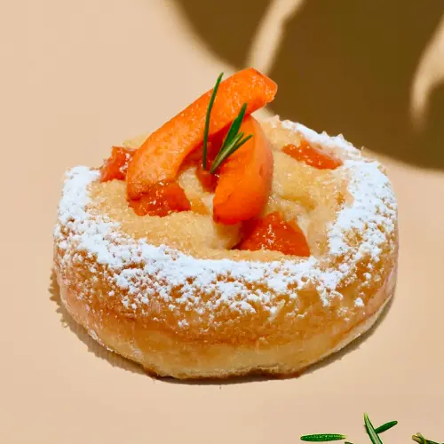 La viennoiserie du mois aux abricots et romarin sur fond beige avec une ombre de feuille et des brindilles de romarins éparpillées