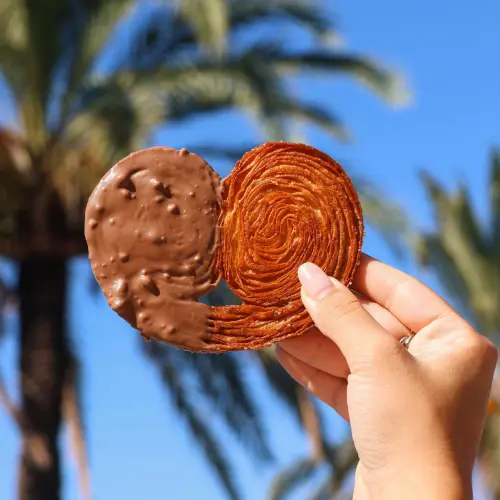 ⁠Une main qui tient le gâteau « palmier rocher » sur la promenade des anglais avec les vrais palmiers en fond