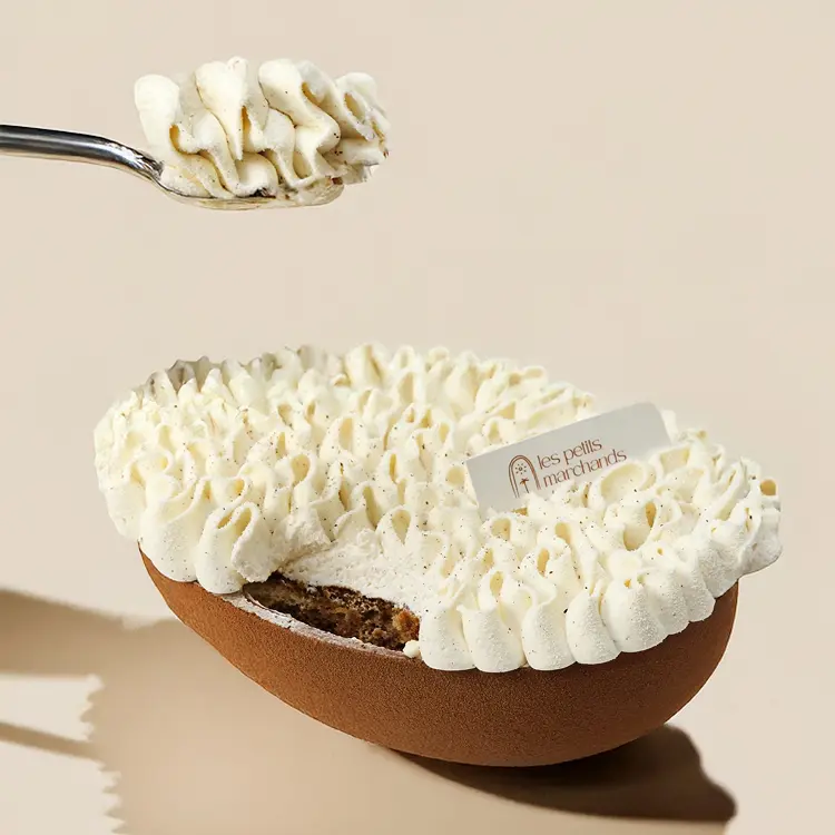 Tiramisu niché dans un demi oeuf au chocolat noir avec une main qui a pris une cuillère, sur fond beige