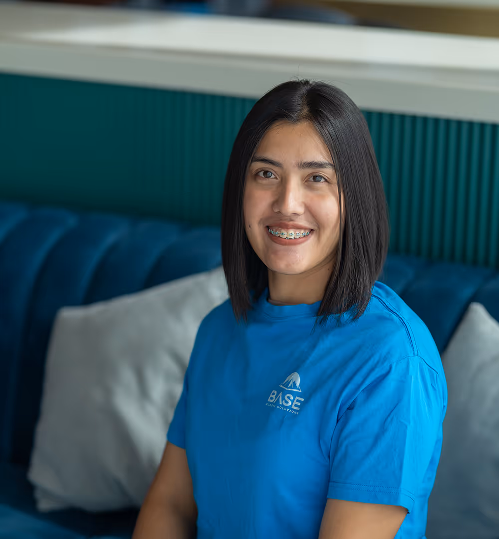 Smiling woman with straight shoulder-length hair wearing a blue BASE Global Solutions t-shirt, sitting on a blue couch with pillows in the background.